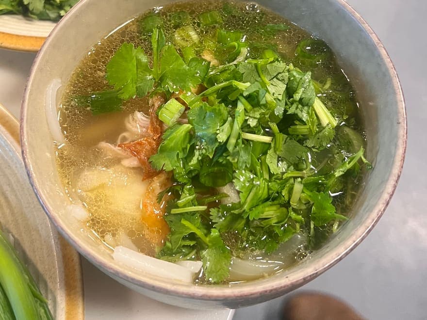A bowl of clear Lao broth heaped with coriander, spring onion and lemongrass.